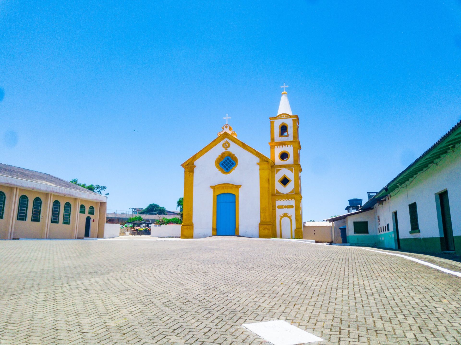Fachada da Capela São João Batista, em branco e amarelo, com portão azul e torre sineira
