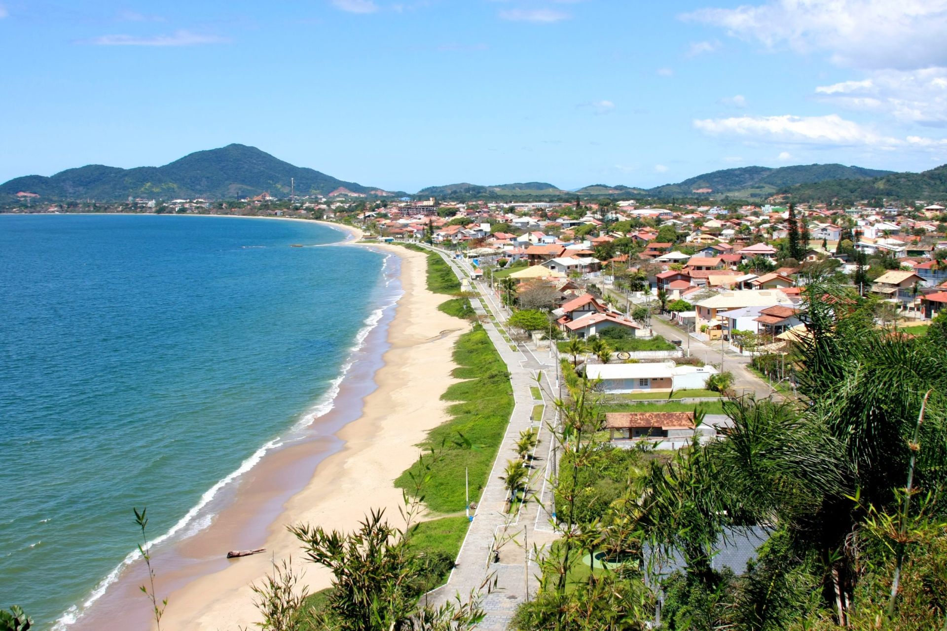 Vista da Praia do Quilombo, em Penha, com longa faixa de areia, orla arborizada e morros ao fundo