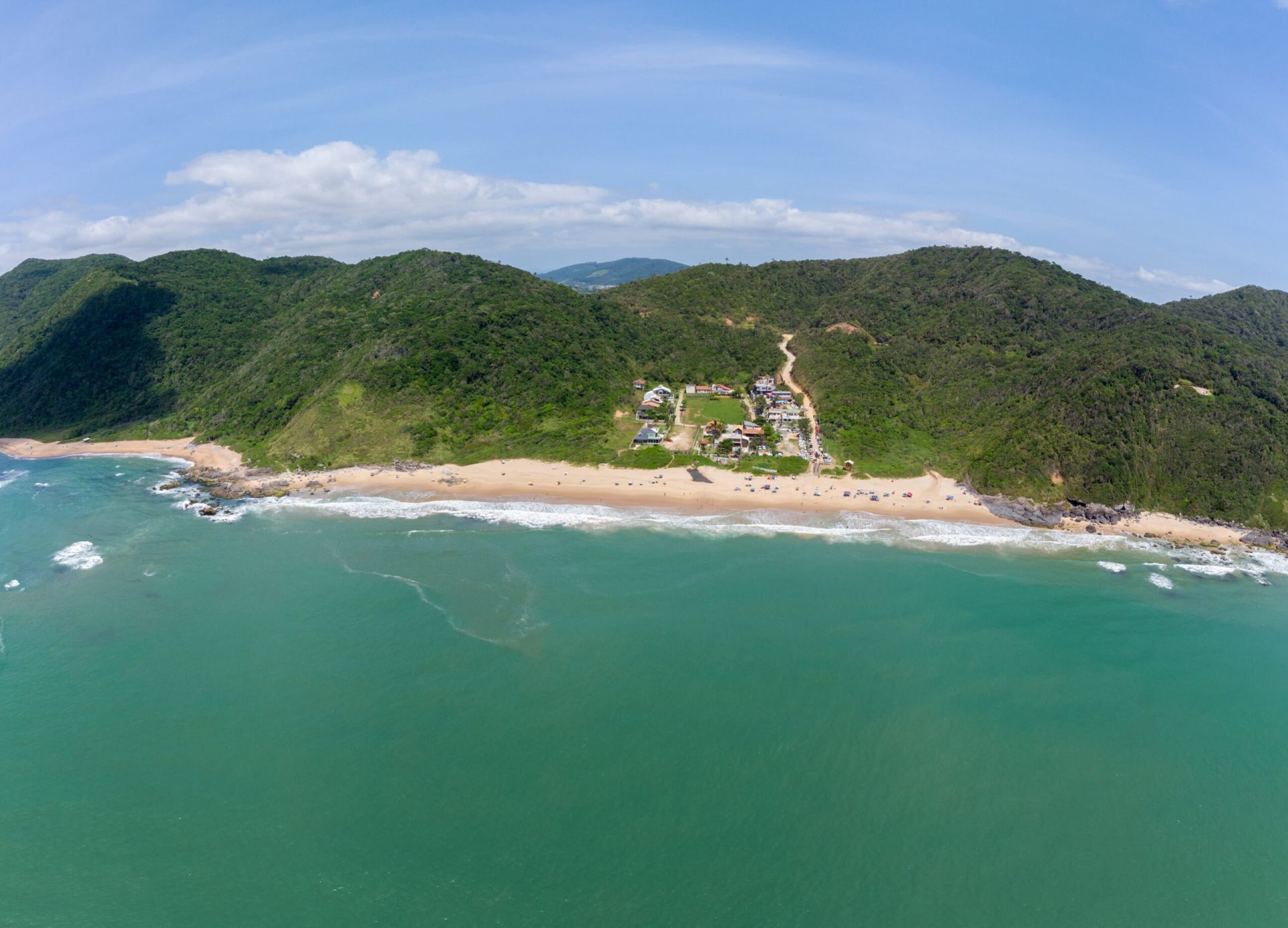 Vista aérea da Praia Vermelha, em Penha, cercada por morros de mata atlântica e mar esverdeado