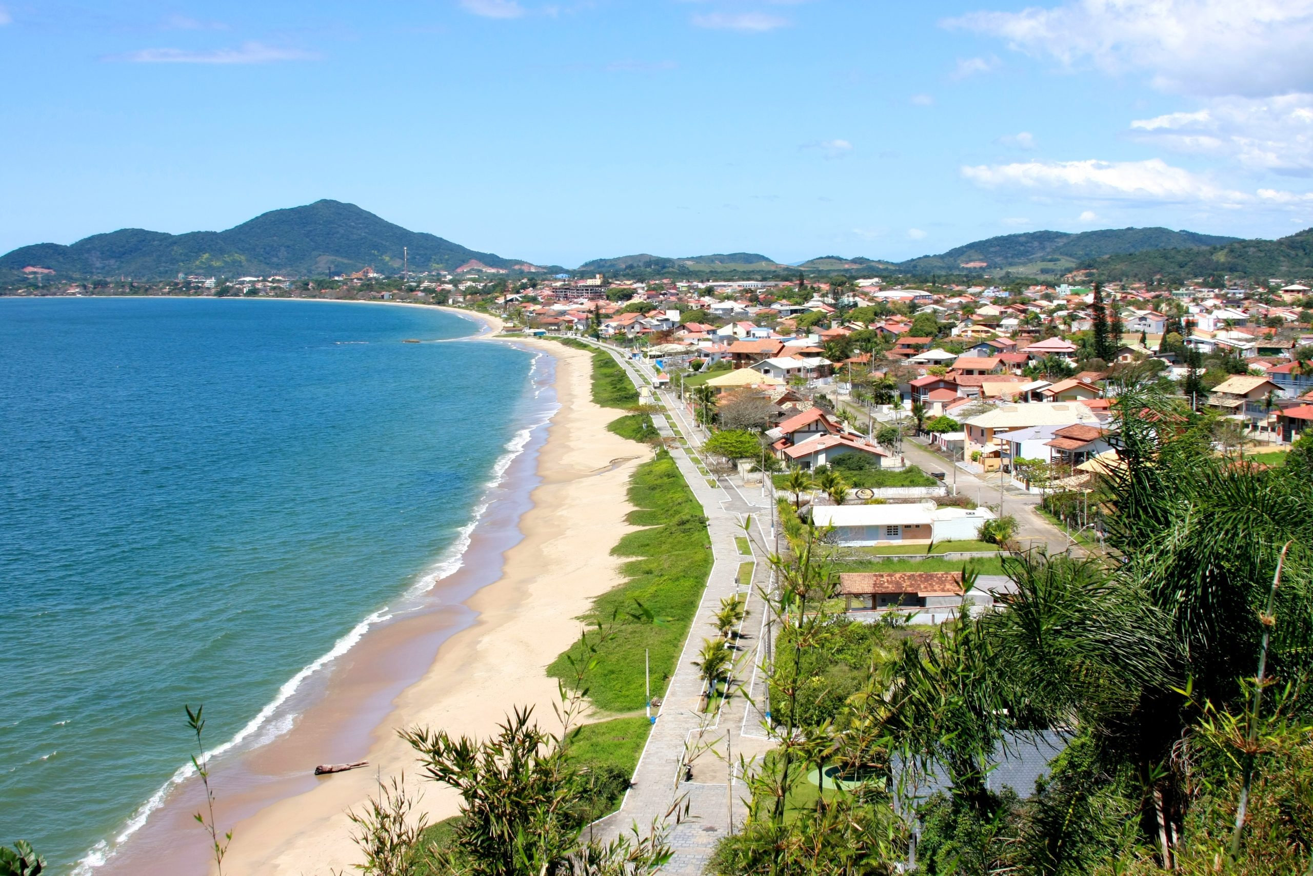 Vista da Praia do Quilombo, em Penha, com longa faixa de areia, orla arborizada e morros ao fundo