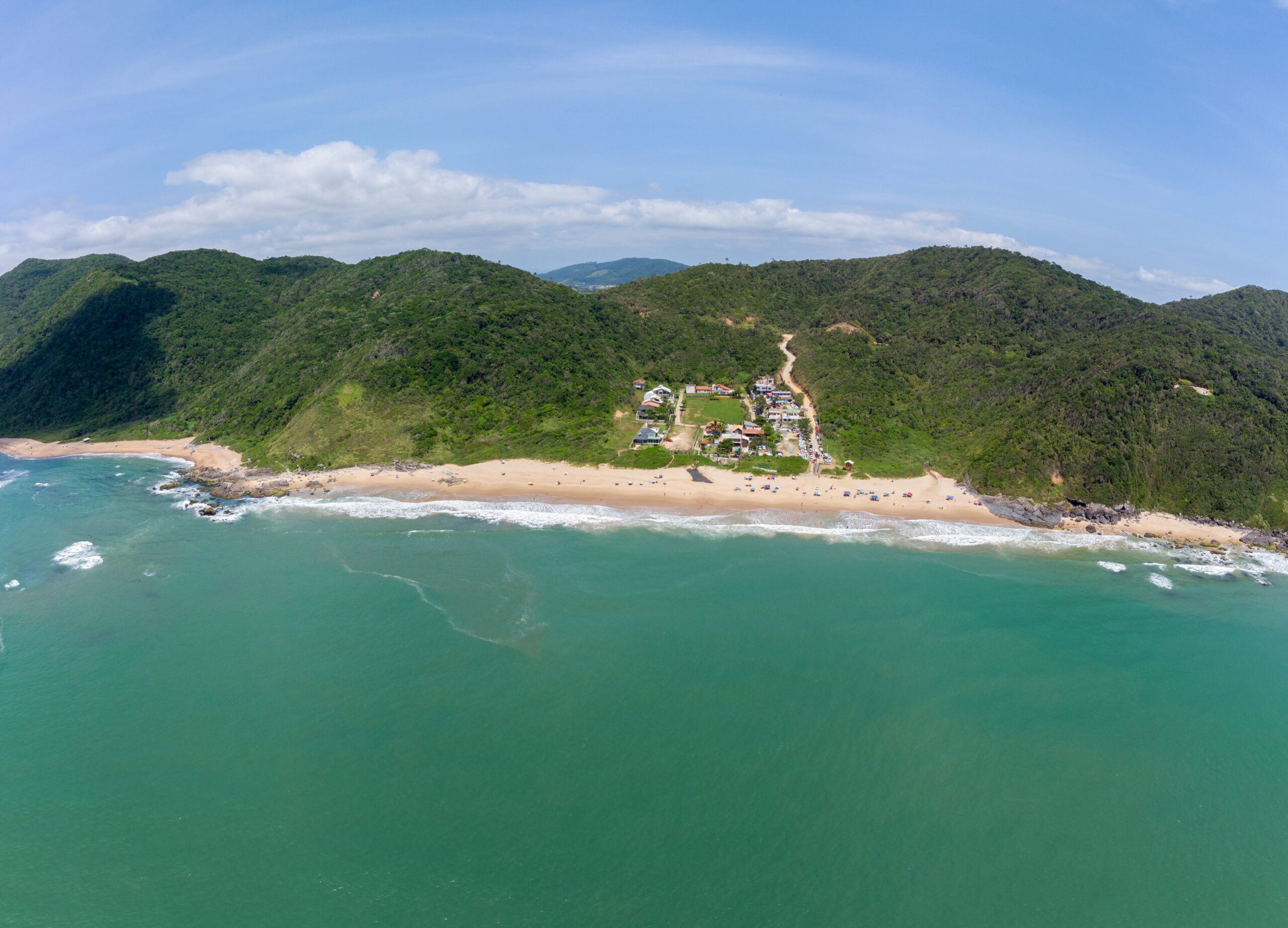 Vista aérea da Praia Vermelha, em Penha, cercada por morros de mata atlântica e mar esverdeado